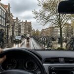 View from inside a car driving through a street in Amsterdam with canal houses, bicycles, and a canal in the background.