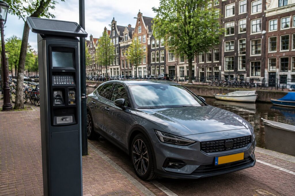 A modern car parked near a roadside payment machine on a street in Amsterdam with traditional buildings, bicycles, and a canal in the background.