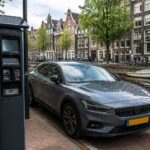 A modern car parked near a roadside payment machine on a street in Amsterdam with traditional buildings, bicycles, and a canal in the background.