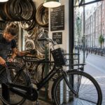 A bike repair shop in an Amsterdam neighborhood with a mechanic fixing a bicycle and colorful buildings visible outside.