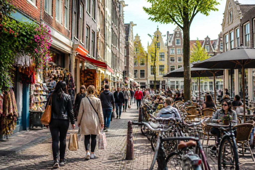 A busy Amsterdam street with historic buildings, people shopping, outdoor cafés, and bicycles parked along the cobblestone road.