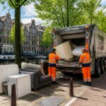 A street in Amsterdam with bulky trash items on the sidewalk and workers loading them into a garbage truck near canal houses.