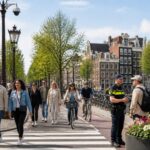 People walking and cycling safely along a canal in Amsterdam with classic buildings and a police officer talking to a tourist.