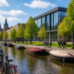 University campus in Amsterdam with modern buildings, students walking, bicycles parked, and a canal nearby under a clear sky.