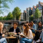A group of students sitting and talking by a canal in Amsterdam with bicycles and traditional houses in the background.