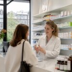 A pharmacist assisting a customer inside a bright, well-organized pharmacy with shelves of medications and large windows showing a city view.