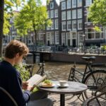 A person sitting alone at an outdoor café table by a canal in Amsterdam, reading a book and having lunch.