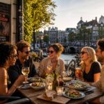 People dining outdoors at a café by a canal in Amsterdam with traditional buildings in the background.