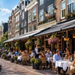 Street view of Dutch canal houses with outdoor seating at restaurants and people dining on a sunny day.
