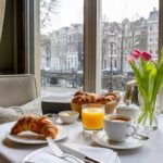 Breakfast table set with croissants, coffee, and orange juice inside a hotel with a view of Amsterdam canals and buildings through large windows.