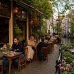 Outdoor seating area of a cozy Amsterdam restaurant by a canal with people dining and traditional buildings in the background.