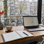 A workspace by a window showing a canal and Amsterdam buildings outside, with a notebook, laptop, coffee cup, and tulips on the desk.