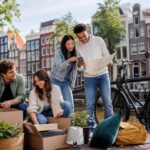 A group of young people unpacking boxes and looking at a map near a canal with colorful houses and a bicycle in Amsterdam.