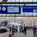 A busy Amsterdam train station with trains, travelers carrying luggage and bicycles, and Dutch-style buildings in the background.