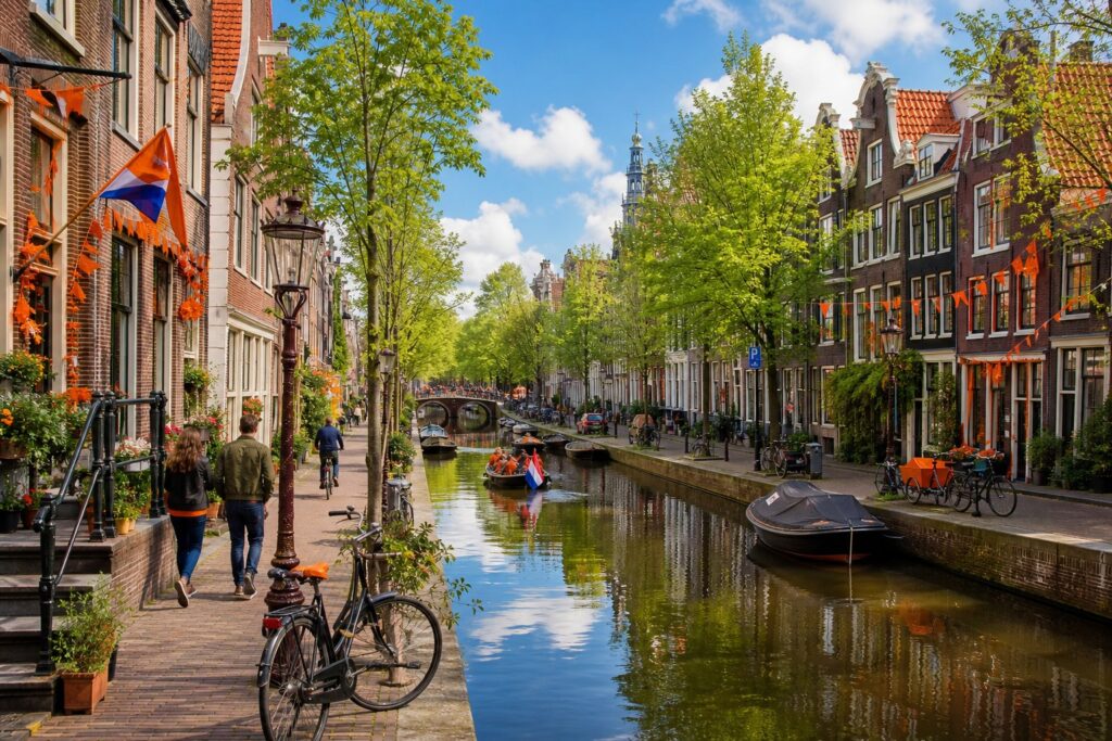 A peaceful Amsterdam canal with traditional houses, trees, and people walking and cycling along the water on a clear day.