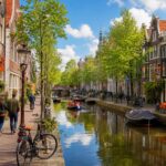 A peaceful Amsterdam canal with traditional houses, trees, and people walking and cycling along the water on a clear day.