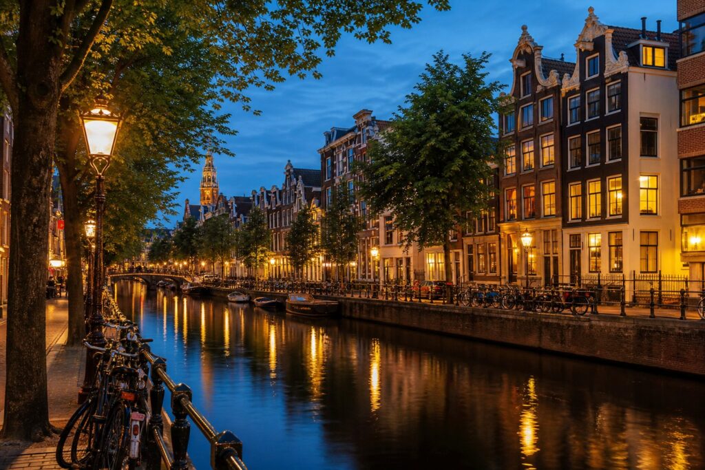 A calm evening scene along an Amsterdam canal with traditional houses, a cobblestone walkway, bicycles, and street lamps reflecting on the water.