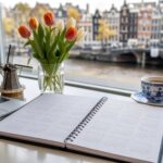 A desk with an open calendar showing months of 2026 and 2027, decorated with tulips, a small windmill model, and a laptop, with a blurred view of Amsterdam buildings and canals outside the window.