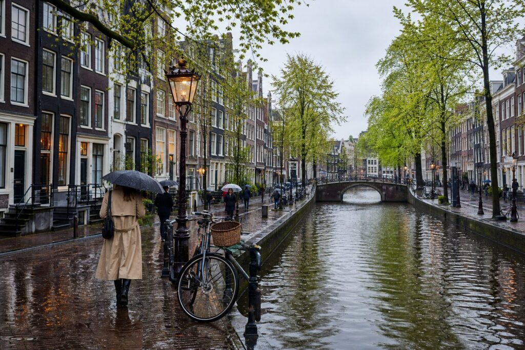 A rainy day in Amsterdam showing canal houses, a wet cobblestone street, people with umbrellas, a bicycle, and a bridge over the canal.