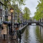 A rainy day in Amsterdam showing canal houses, a wet cobblestone street, people with umbrellas, a bicycle, and a bridge over the canal.