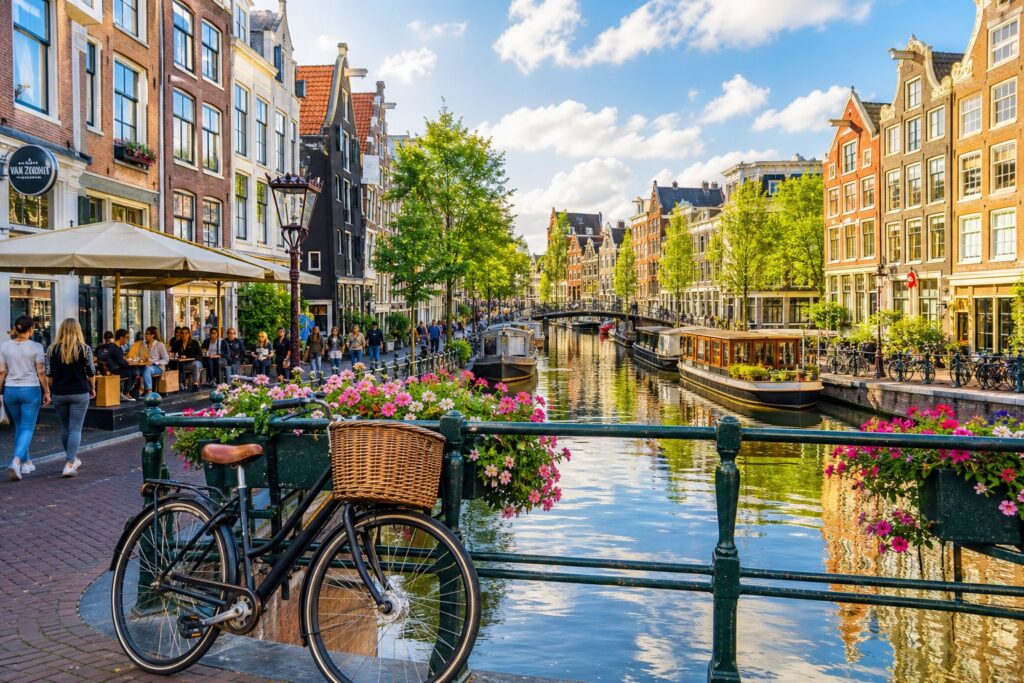 A canal in Amsterdam lined with colorful houses, a bicycle near a flower-adorned bridge, and people walking along the street.