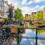 A canal in Amsterdam lined with colorful houses, a bicycle near a flower-adorned bridge, and people walking along the street.