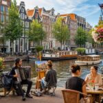 People enjoying outdoor café and street music along a canal with traditional Dutch houses in Amsterdam.