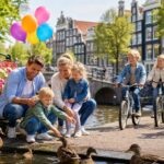 A family with children enjoying a sunny day by a canal in Amsterdam with bicycles, a wooden bridge, and traditional houses in the background.