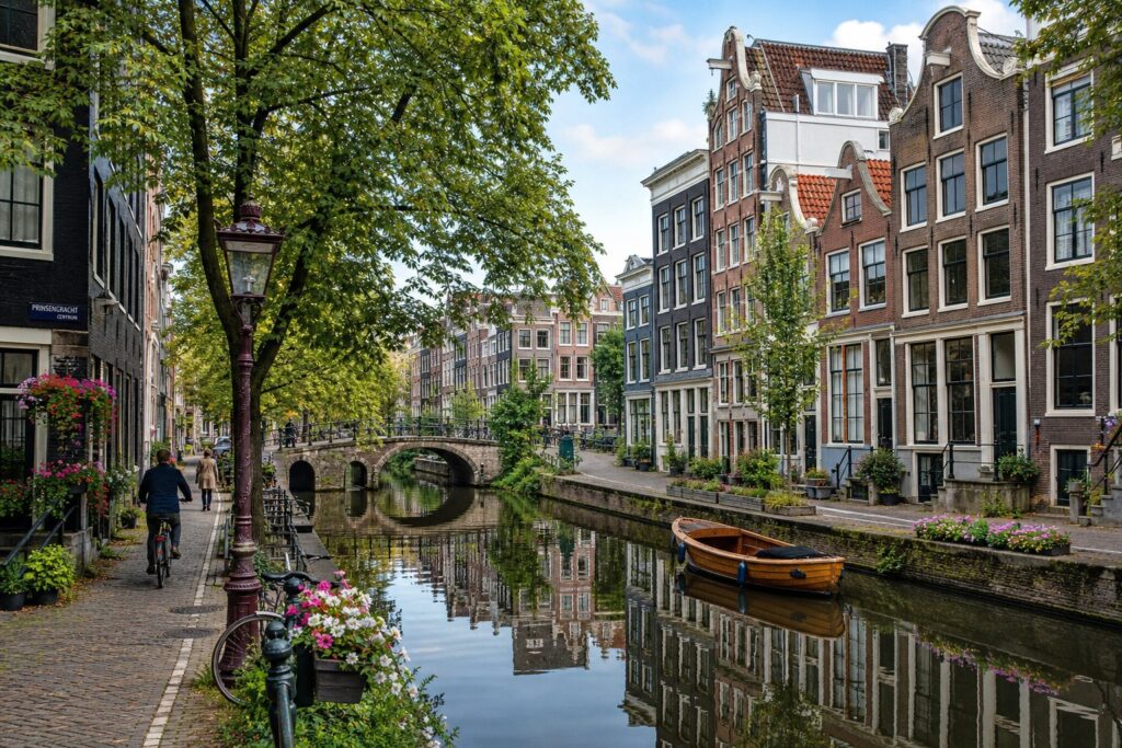 A quiet canal in Amsterdam with traditional houses, trees, and a small boat, with a few people walking and cycling nearby.