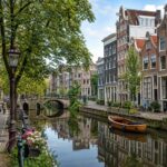 A quiet canal in Amsterdam with traditional houses, trees, and a small boat, with a few people walking and cycling nearby.
