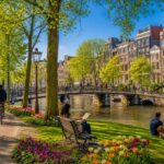 A sunny park in Amsterdam with green trees, colorful flowers, a canal with a pedestrian bridge, and people walking, sitting, and cycling.