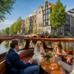 A private saloon boat cruising through Amsterdam canals with historic buildings and bridges in the background.