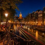 Nighttime view of Amsterdam canals with illuminated historic buildings, outdoor terraces with people, and bicycles along the water.