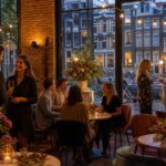 Interior of a small celebration venue in Amsterdam with warm lighting, exposed brick walls, and windows showing canal houses outside. People are enjoying a relaxed gathering with drinks.