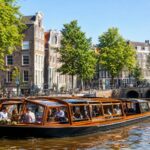 A modern boat cruising along an Amsterdam canal with historic buildings and green trees lining the water.