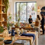 Interior of an Amsterdam design shop with shelves and tables displaying handcrafted home decor and artisanal products, with a local maker assisting customers.