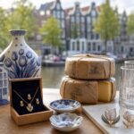 A display of elegant Amsterdam-inspired souvenirs including Delft blue pottery, tulip-themed jewelry, Dutch cheese, and glassware with canal house designs on a wooden table with canal houses visible in the background.