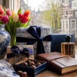 A table near a window displaying elegant wrapped gifts, tulips, a porcelain vase, chocolates, and perfume with a blurred view of Amsterdam canal houses outside.