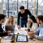 A group of young professionals collaborating around a table with laptops and documents in an office with windows showing Amsterdam canal houses outside.