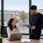 A group of young professionals working and discussing in a modern office with a view of Amsterdam canals and traditional buildings.
