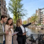 A young couple discussing housing options with a real estate agent near a canal in Amsterdam, with traditional Dutch houses and bicycles in the background.