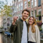A young couple smiling and holding keys in front of a traditional Amsterdam canal house with boats and bicycles nearby.
