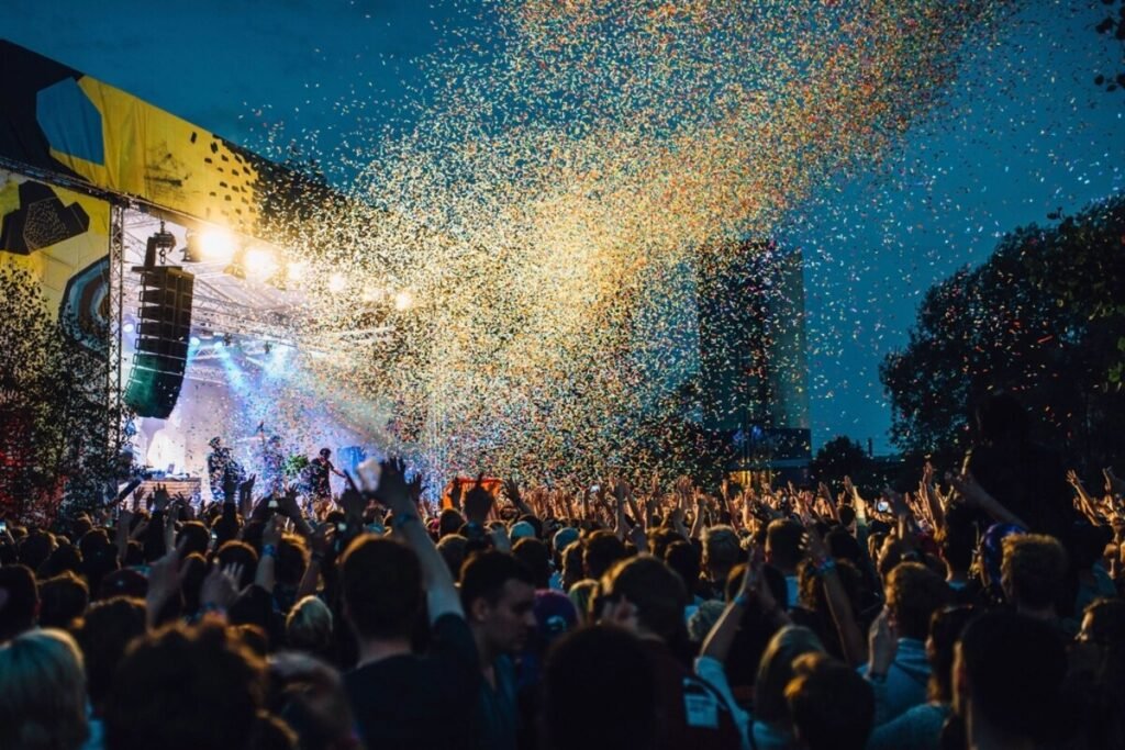 A crowd with hands raised in front of an outdoor festival stage as confetti falls