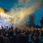 A crowd with hands raised in front of an outdoor festival stage as confetti falls