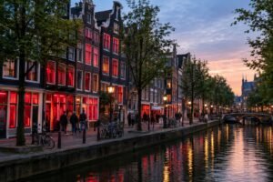 A canal-side street in Amsterdam with historic buildings, red-lit windows, bicycles, and pedestrians.