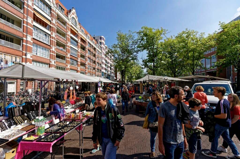 Shoppers walk between the stalls of the Waterlooplein flea market in Amsterdam on a sunny day.